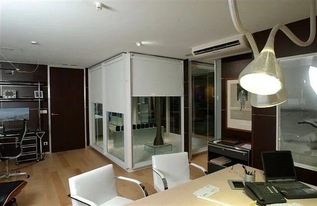 Oficina moderna con sala de paredes de cristal, sillas blancas, escritorios y lámparas colgantes.