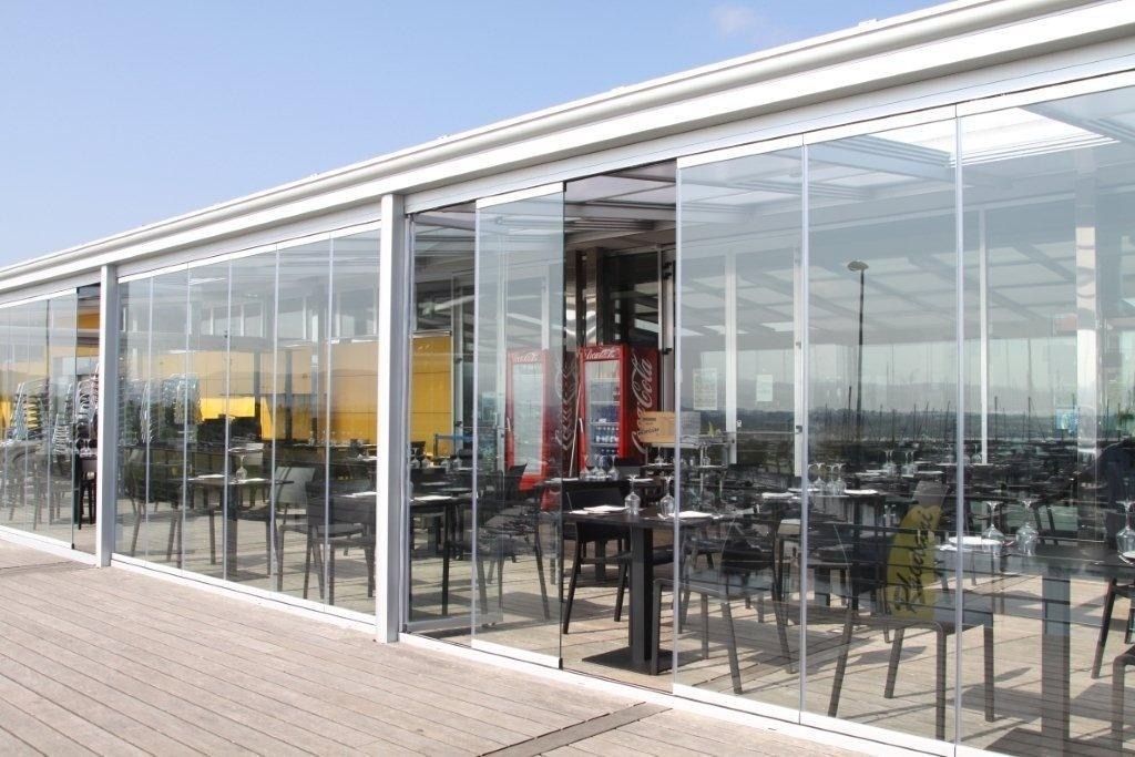 Terraza-cafetería al aire libre con paredes de cristal, mesas y sillas en un paseo pavimentado.