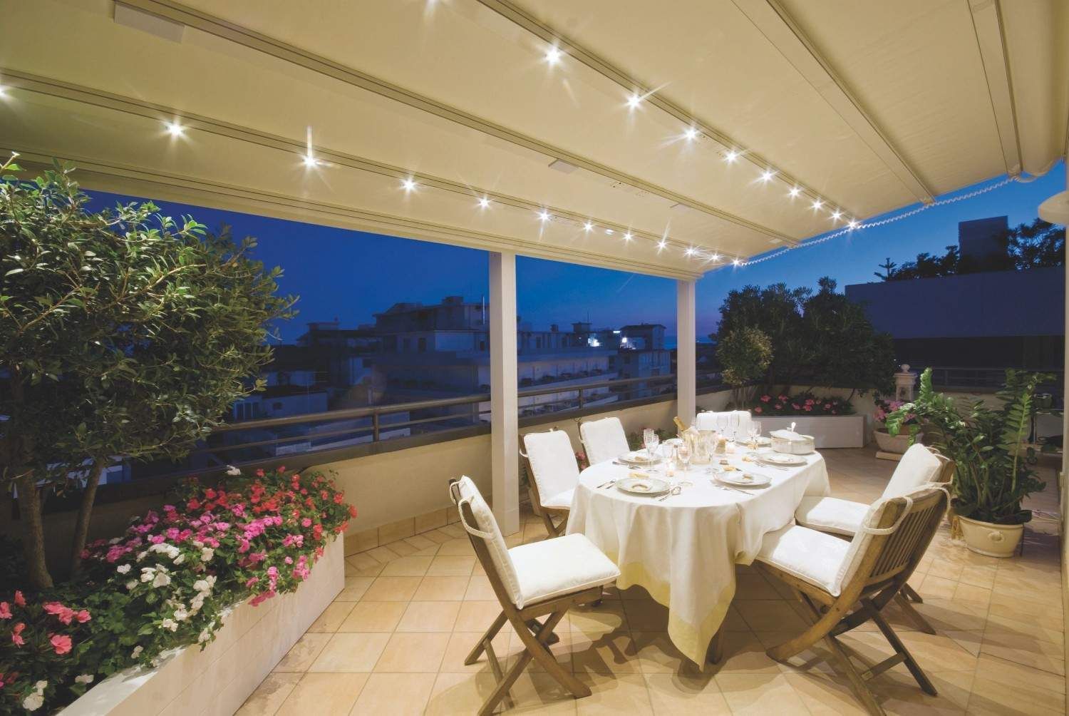 Comedor cubierto en el patio con mesa, sillas blancas, plantas en macetas y luces de guirnalda cálidas al anochecer.