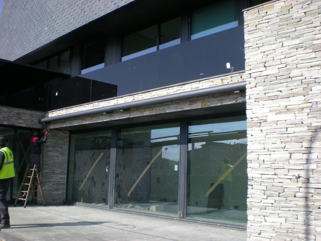 Entrada moderna al edificio con grandes puertas de cristal, fachada de piedra y dos trabajadores cerca de una escalera.