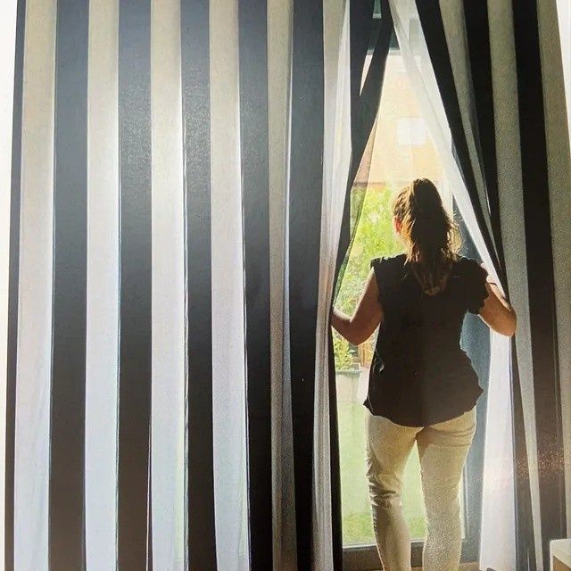 Una persona está de pie junto a unas cortinas a rayas y mira por una ventana luminosa.