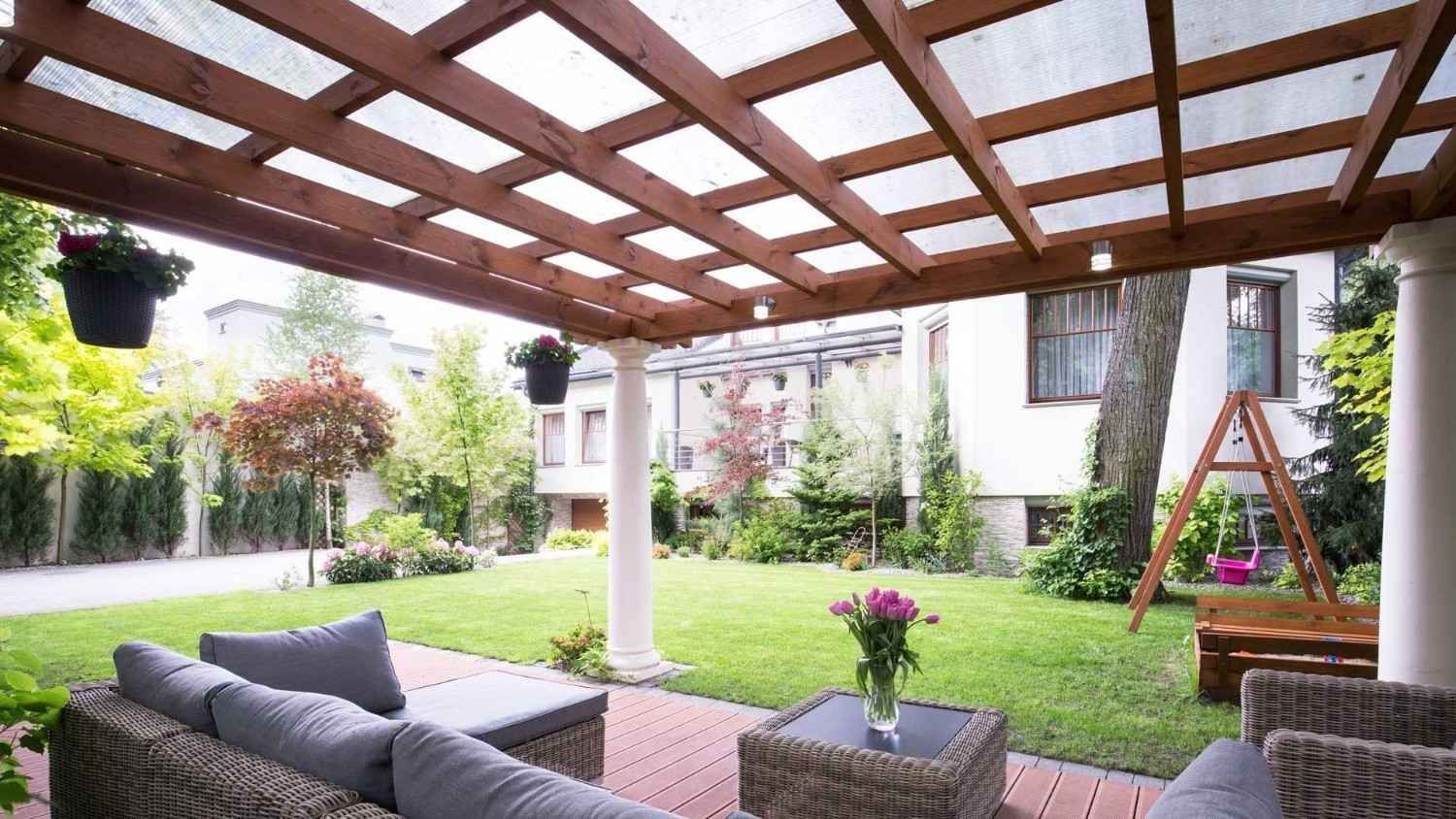 Patio cubierto con asientos de mimbre, plantas colgantes, césped y un columpio de madera en un luminoso jardín trasero.