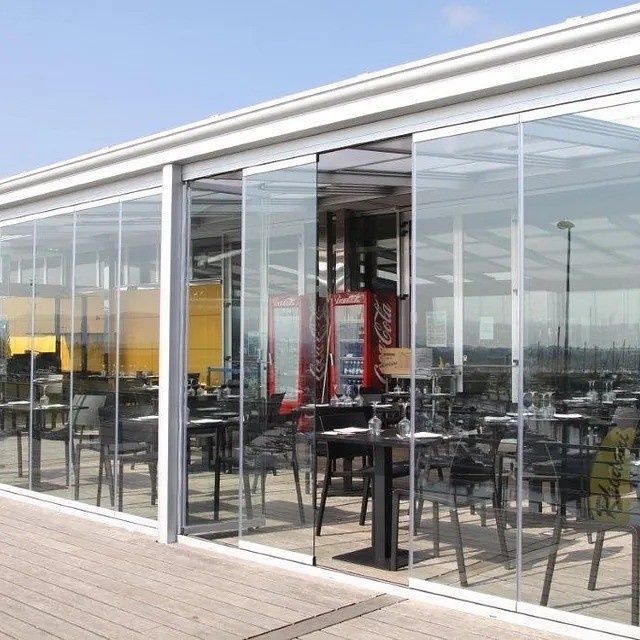 Terraza de cafetería con paredes de cristal, mesas y sillas negras bajo un techo blanco, con vistas al agua.