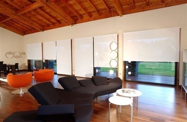 Sala de estar moderna con sofá seccional negro, sillas naranjas, mesas blancas y grandes ventanales.