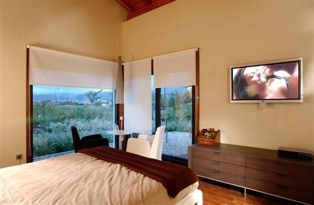 Dormitorio con grandes ventanales, cama, cómoda y televisor montado en la pared con vistas a un paisaje verde.