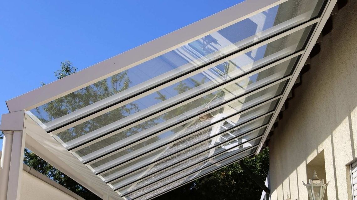 Pérgola blanca con paneles inclinados transparentes junto a una casa beige bajo un cielo azul.