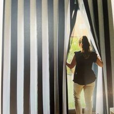 Una persona aparta unas cortinas a rayas y mira a través de una puerta hacia una vista de un verde brillante.
