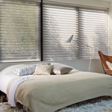 Dormitorio minimalista con cama hecha, almohadas blancas, lámpara de pie y persianas grandes en la ventana.
