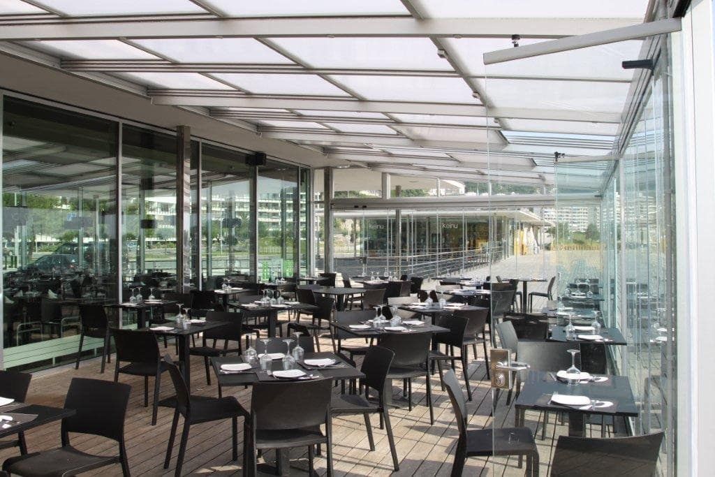 Terraza vacía de una cafetería al aire libre con mesas y sillas negras bajo un toldo de cristal.