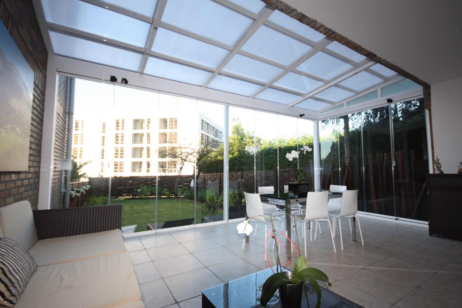 Patio cubierto con mesa de cristal, sillas blancas, sofá y vistas a edificios de apartamentos y zonas verdes.