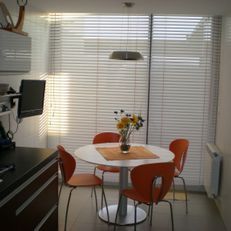 Cocina pequeña con mesa redonda blanca, sillas naranjas, jarrón con flores y persianas en las ventanas.