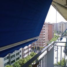 Balcón con toldo azul con vistas a edificios de apartamentos y a una calle arbolada.