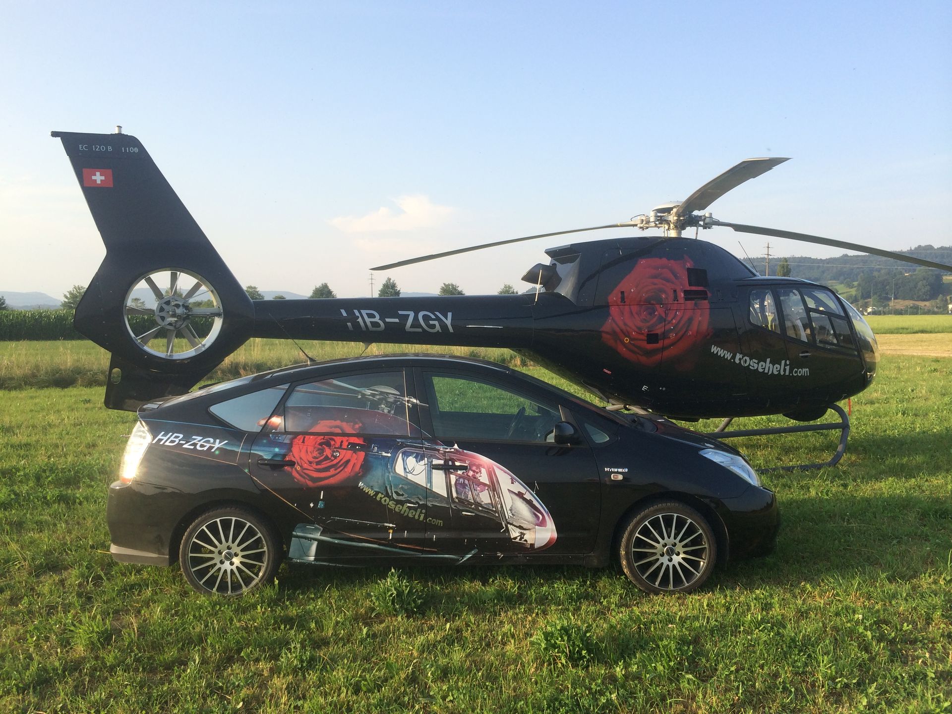 Auf einer Wiese parkt ein schwarzes Auto neben einem Hubschrauber.