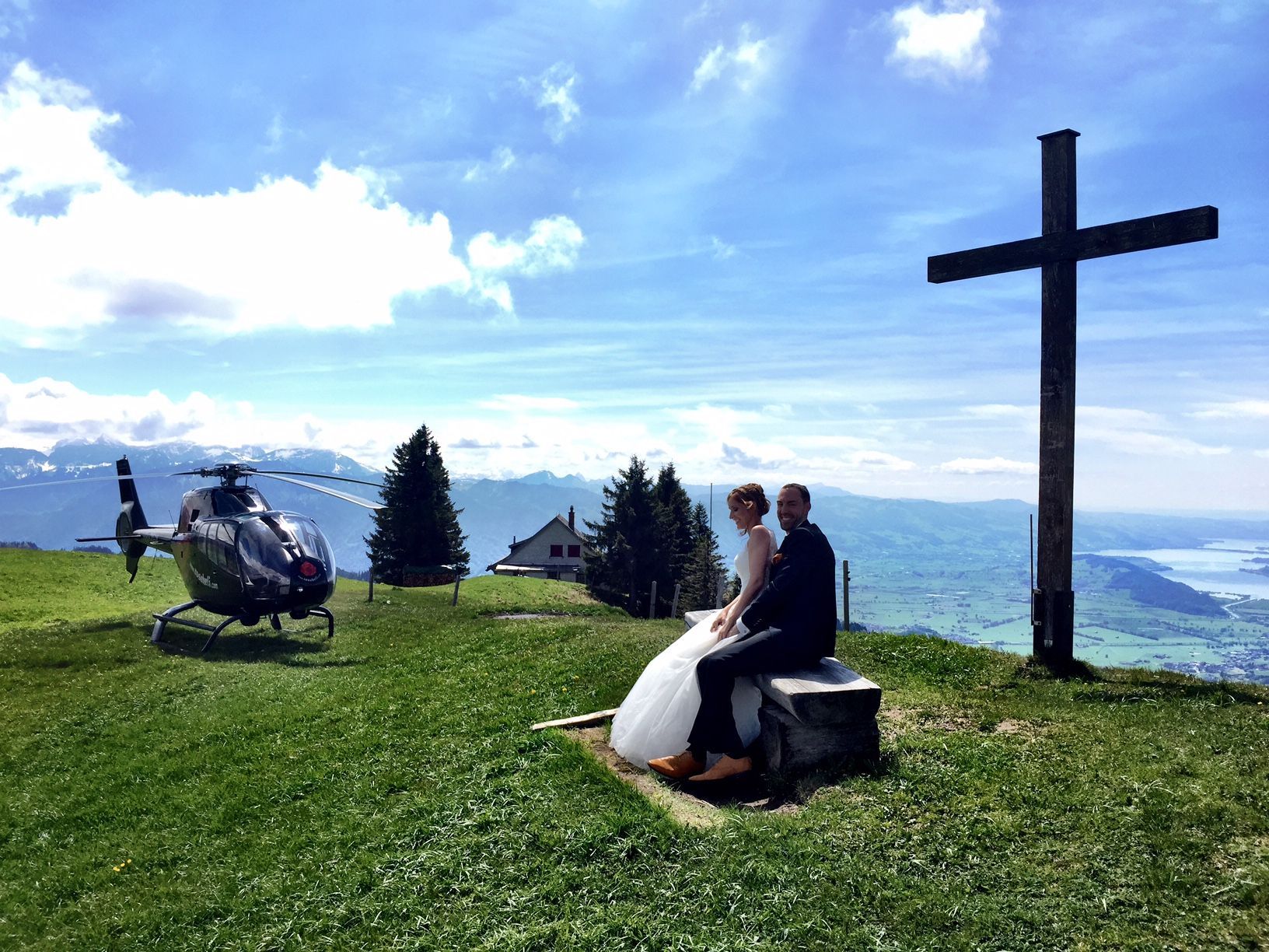 Eine Braut und ein Bräutigam sitzen auf einer Bank neben einem Gipfelkreuz und einem Hubschrauber.