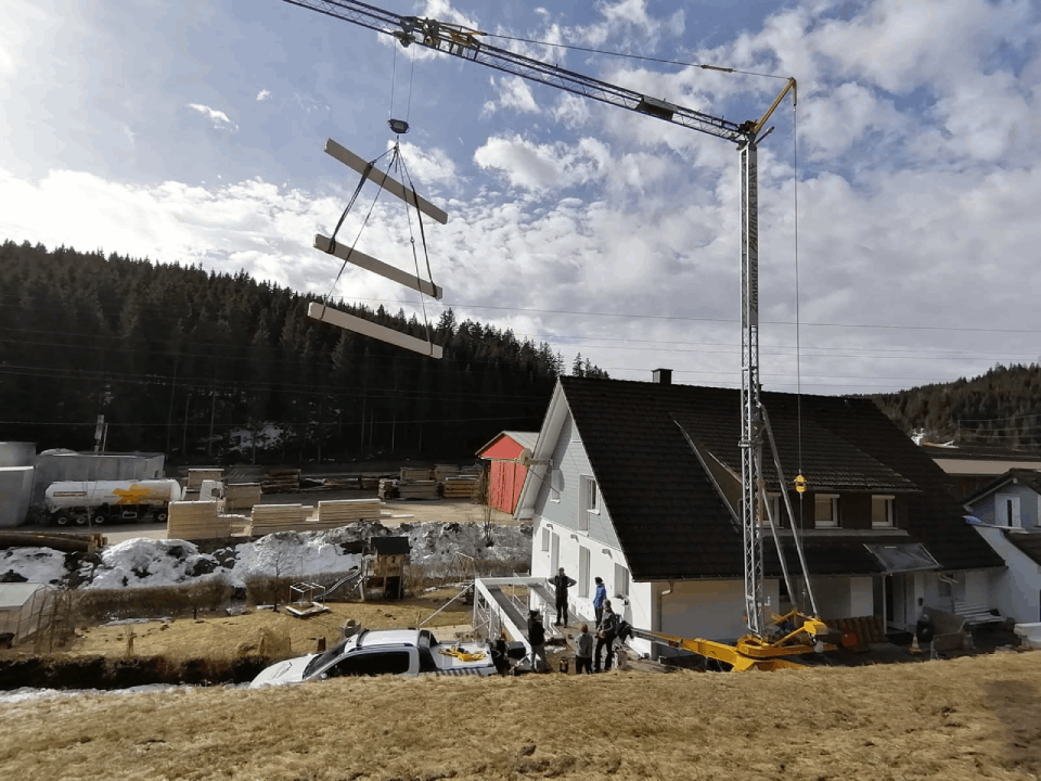 Ein Kran hebt ein Stück Holz vor einem Haus an, Zimmerei Patrick Pfaff