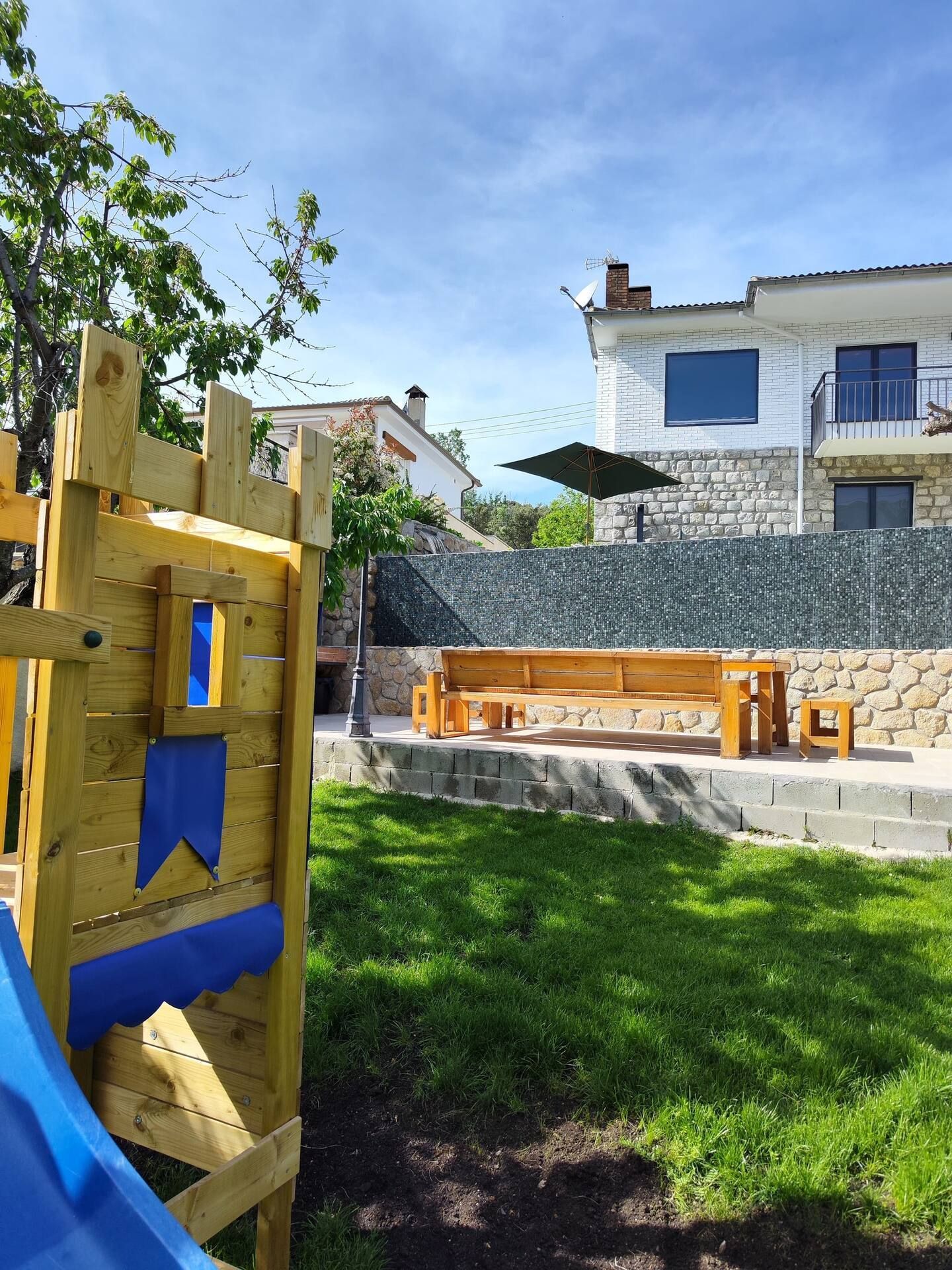 Un parque infantil de madera con tobogán y bancos en un patio trasero con una casa al fondo.