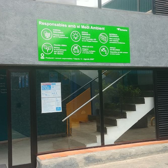 Letrero verde: “Responsable con el Medio Ambiente” sobre la entrada de un edificio, con íconos y texto, escalera en el interior.