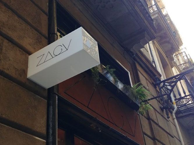 Letrero para tienda ZACV, caja blanca con letras negras, montado en un poste afuera de un edificio marrón.