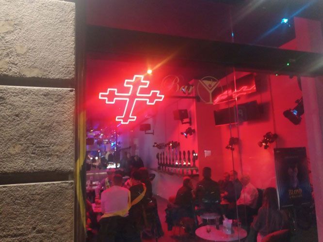 Interior de bar iluminado con una cruz de neón. La gente está sentada en las mesas, charlando.