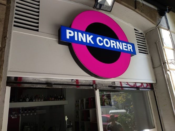 Letrero de bar Pink Corner: círculo rosa, centro negro, texto azul "PINK CORNER" sobre una puerta.
