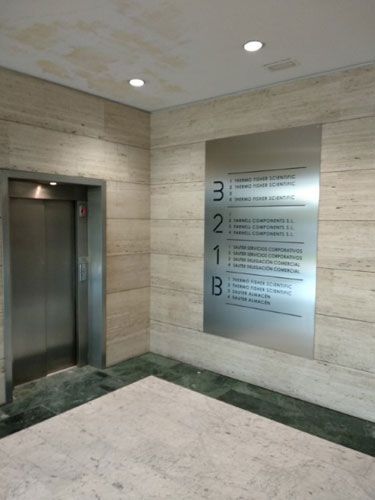 Vestíbulo del ascensor con puerta plateada y un cartel con los pisos y negocios. Paredes beige y suelo verde.
