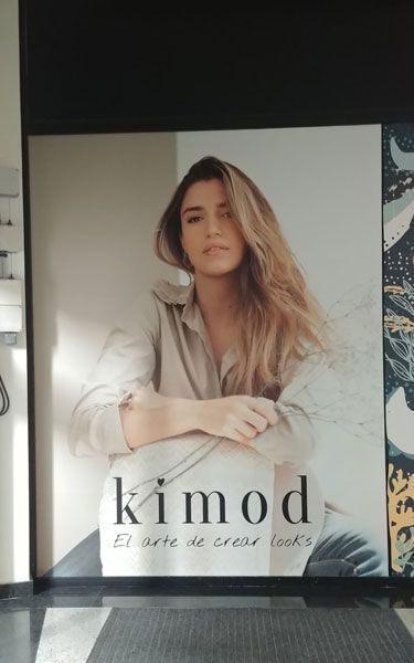 Mujer con camisa beige, anuncio de Kimod. Cabello claro, expresión neutra. Fondo blanco/gris.