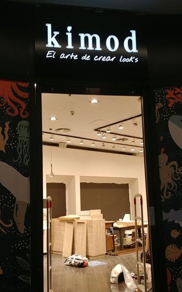 Entrada de la tienda Kimod con un letrero iluminado. El interior tiene materiales de construcción y paredes sin terminar.