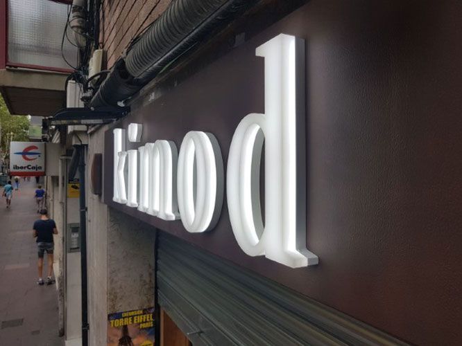 Letrero iluminado "Kinod" en el exterior de un edificio marrón; letras blancas, vista de la calle con peatones.