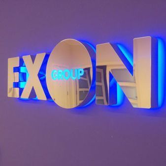 Logotipo metálico “EXON GROUP” con retroiluminación azul sobre una pared violeta.