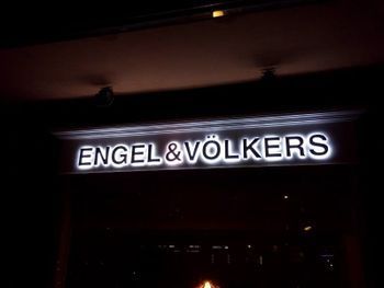 Letrero luminoso: Agencia inmobiliaria Engel & Völkers. Texto blanco sobre fondo oscuro.