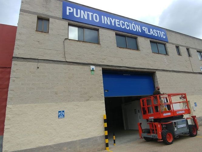 Exterior de nave industrial Punto Inyección Plástico con ascensor frente a puerta azul abierta.