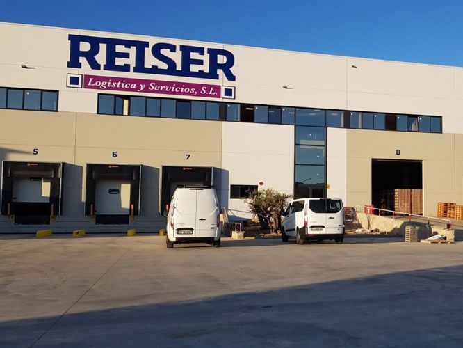 Exterior del almacén de Reiser Logistics. Furgonetas blancas estacionadas en los muelles de carga. Edificio beige con logotipo azul.