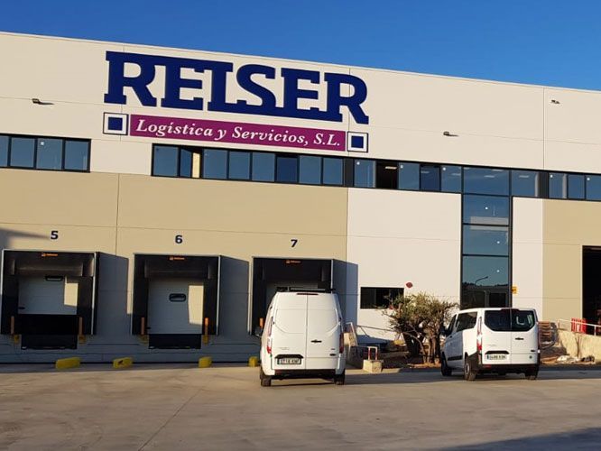 Almacén de REISER Logística y Servicios con furgonetas en los muelles de carga. Edificio azul y blanco.