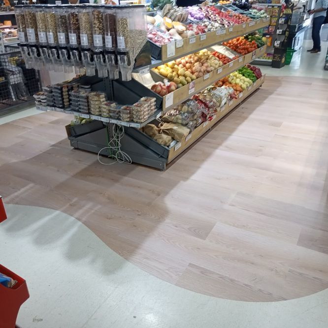 Exhibición de productos en una tienda de comestibles con frutas y verduras; piso con apariencia de madera clara.