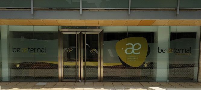 Exterior de una tienda con ventanas y puertas de cristal. El letrero dice "Sé eterno". Un diseño amarillo con "ae" está en la ventana.
