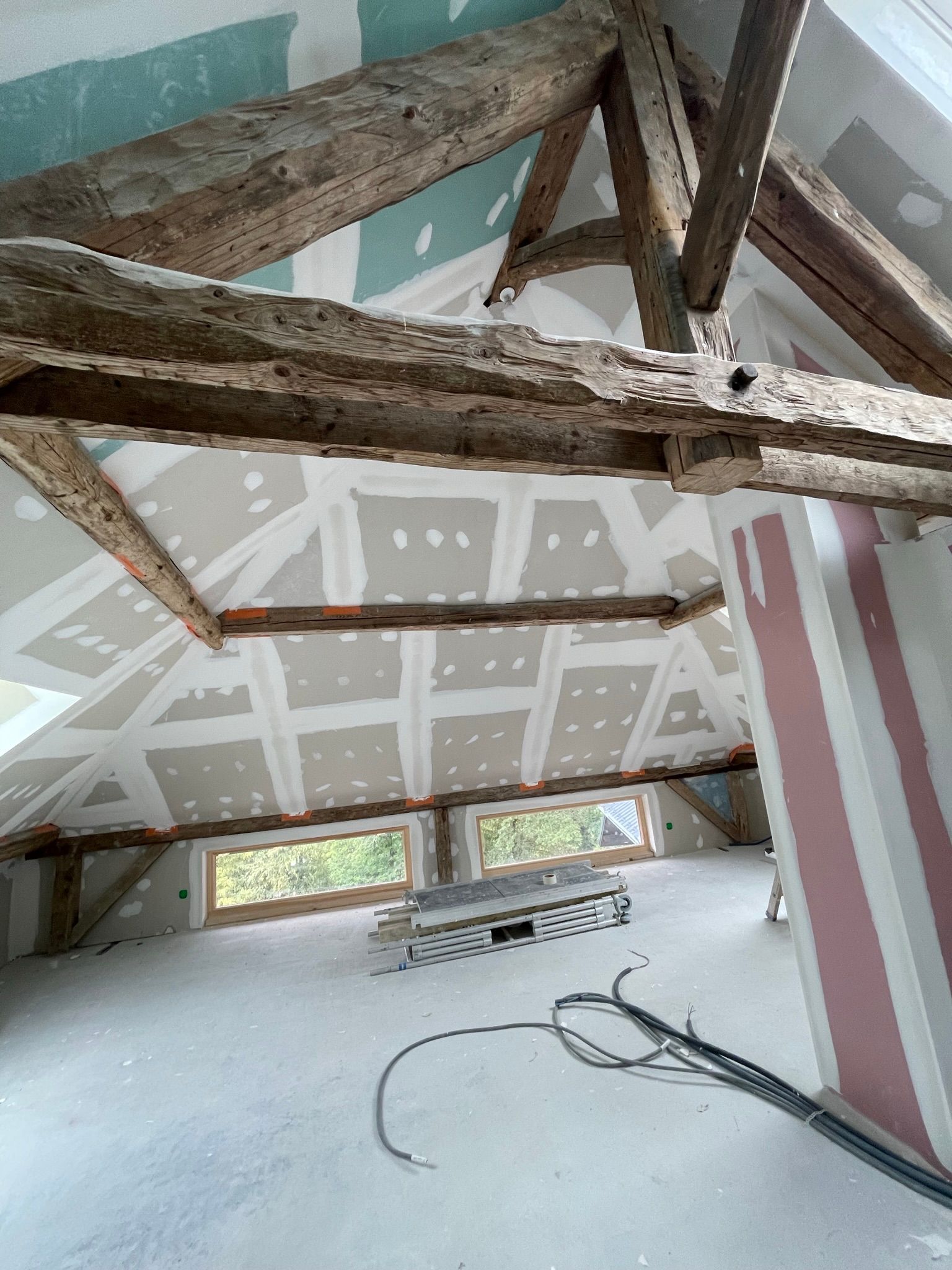 Rénovation de combles avec poutres apparentes, cloisons sèches brutes aux joints collés et fenêtres basses.