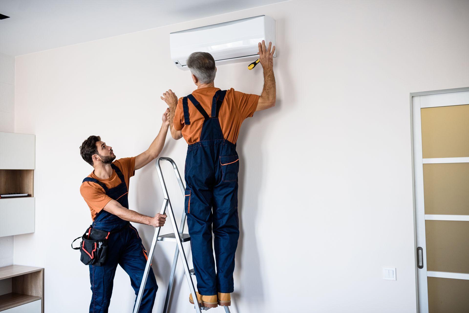 Deux techniciens installent un climatiseur blanc sur un mur.