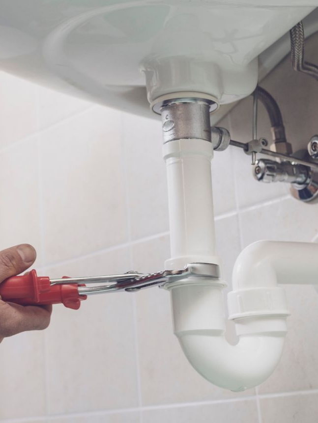Un plombier utilise une clé pour serrer un tuyau en plastique blanc sous un lavabo de salle de bain.