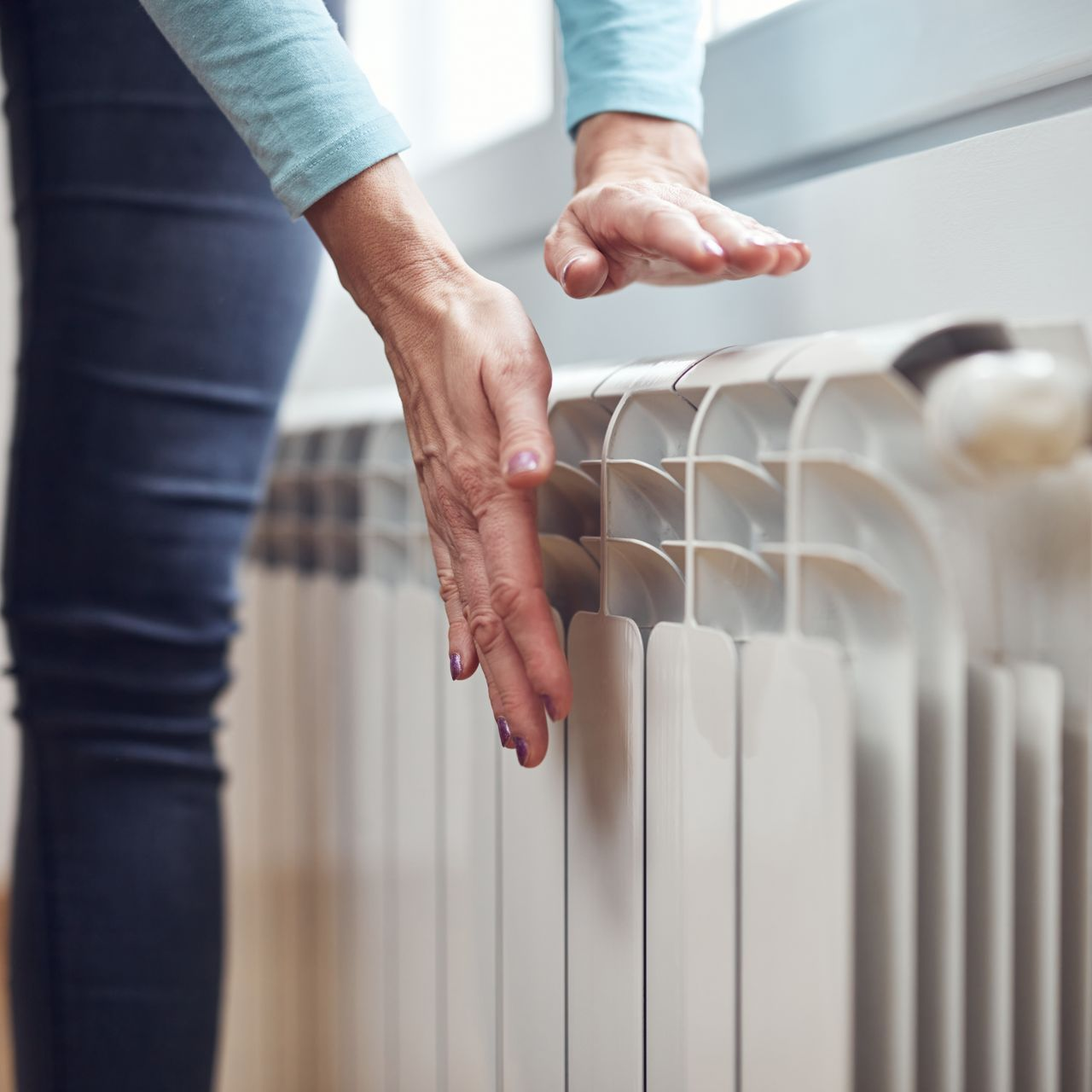Une personne touche un radiateur blanc avec les mains, vérifiant s'il chauffe près d'une fenêtre.