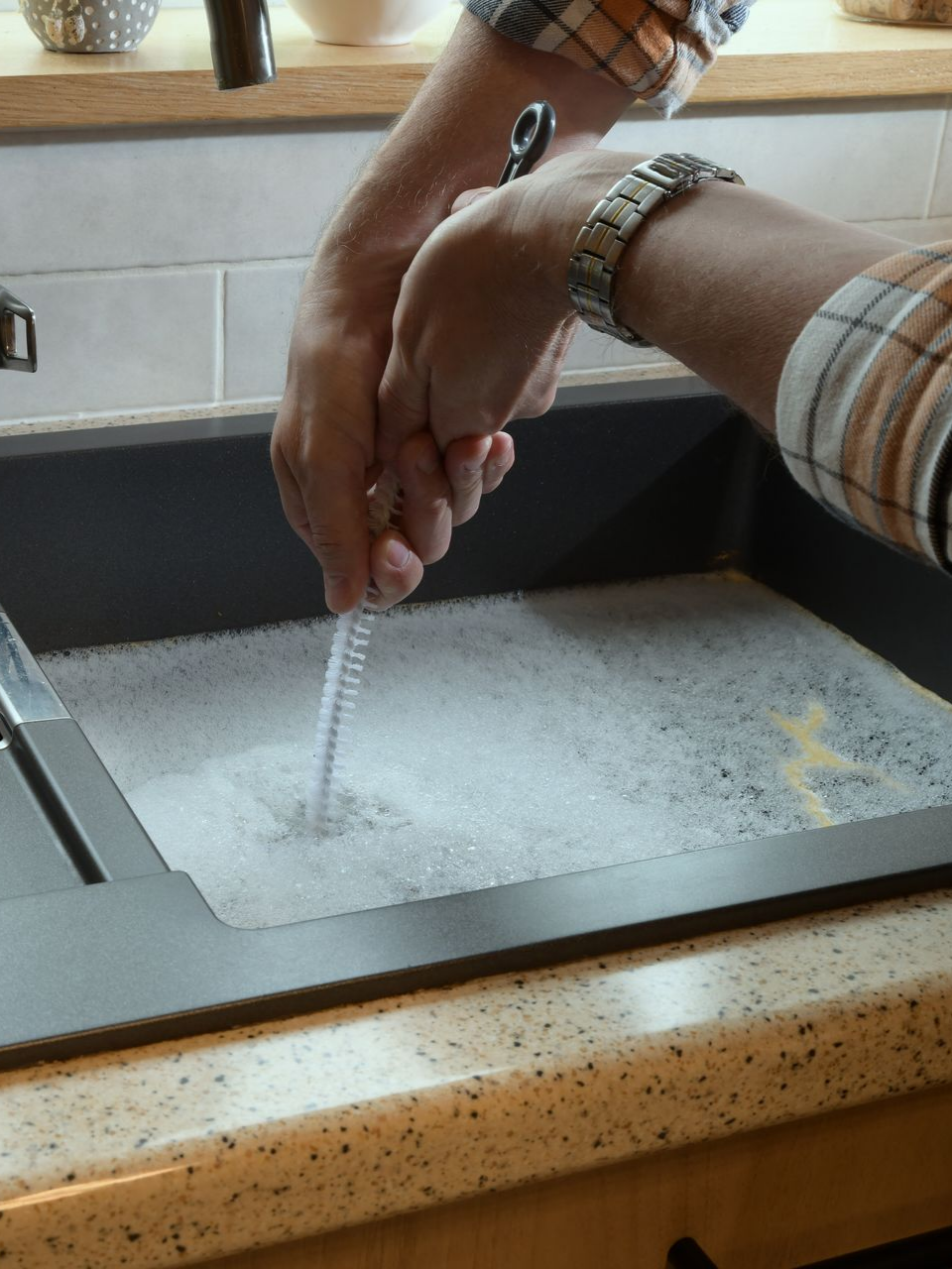 Des mains lavent quelque chose dans un évier de cuisine rempli d'eau savonneuse. On utilise une brosse enroulée.