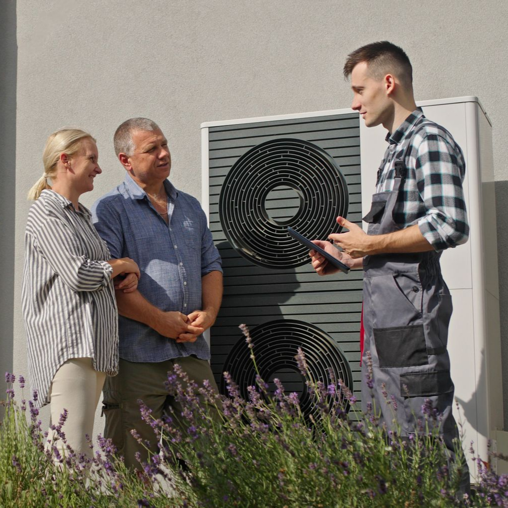 Un technicien explique le fonctionnement d'une pompe à chaleur à un couple devant leur maison.