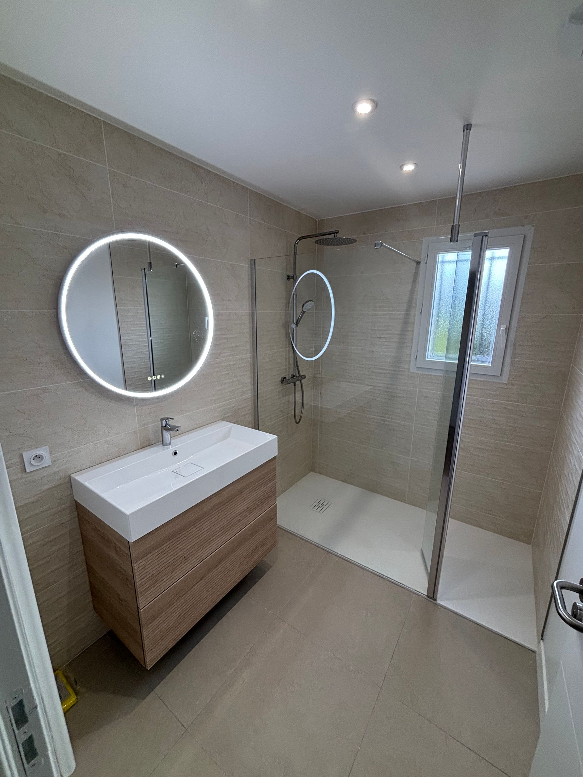 Salle de bains moderne avec meuble vasque en bois, miroir rond rétroéclairé, douche et murs carrelés.