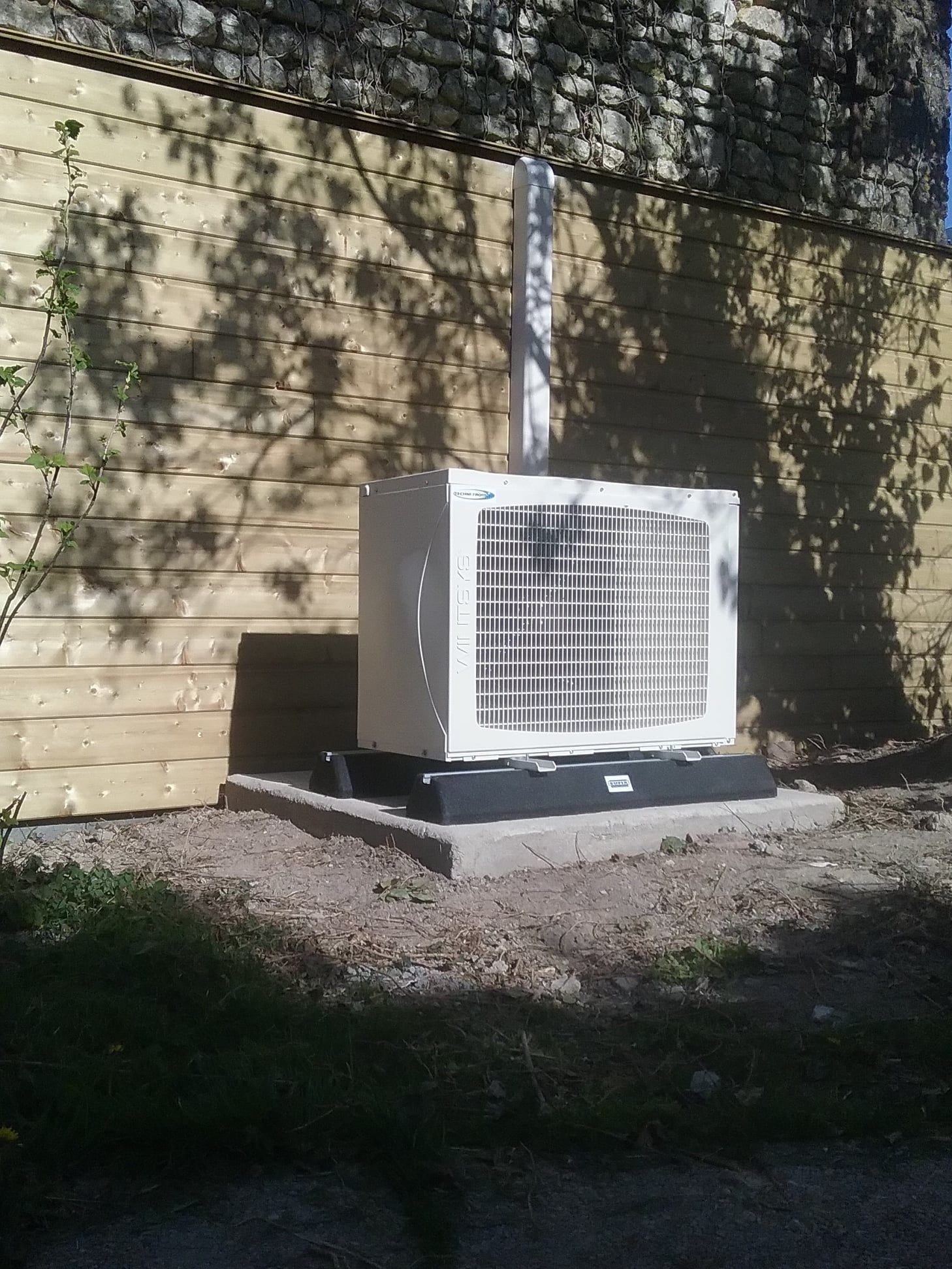 Unité de climatisation blanche à l'extérieur d'un bâtiment, montée sur une base noire contre un mur en bois ; un tuyau vertical s'étend vers le haut.