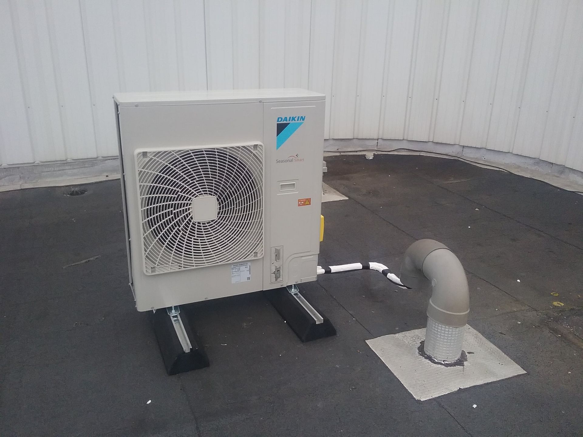 Unité de climatisation de toit beige avec un grand ventilateur, montée sur des supports noirs à côté d'un tuyau d'évacuation sur un toit sombre.