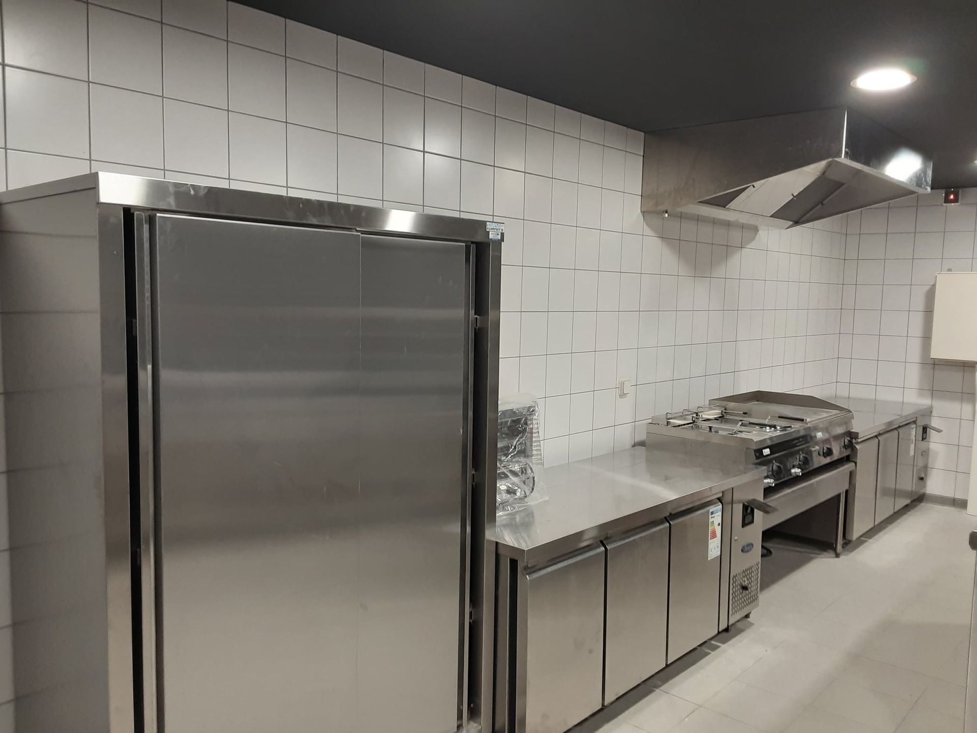 Cuisine commerciale en acier inoxydable avec réfrigérateur, plaque de cuisson et hotte aspirante.