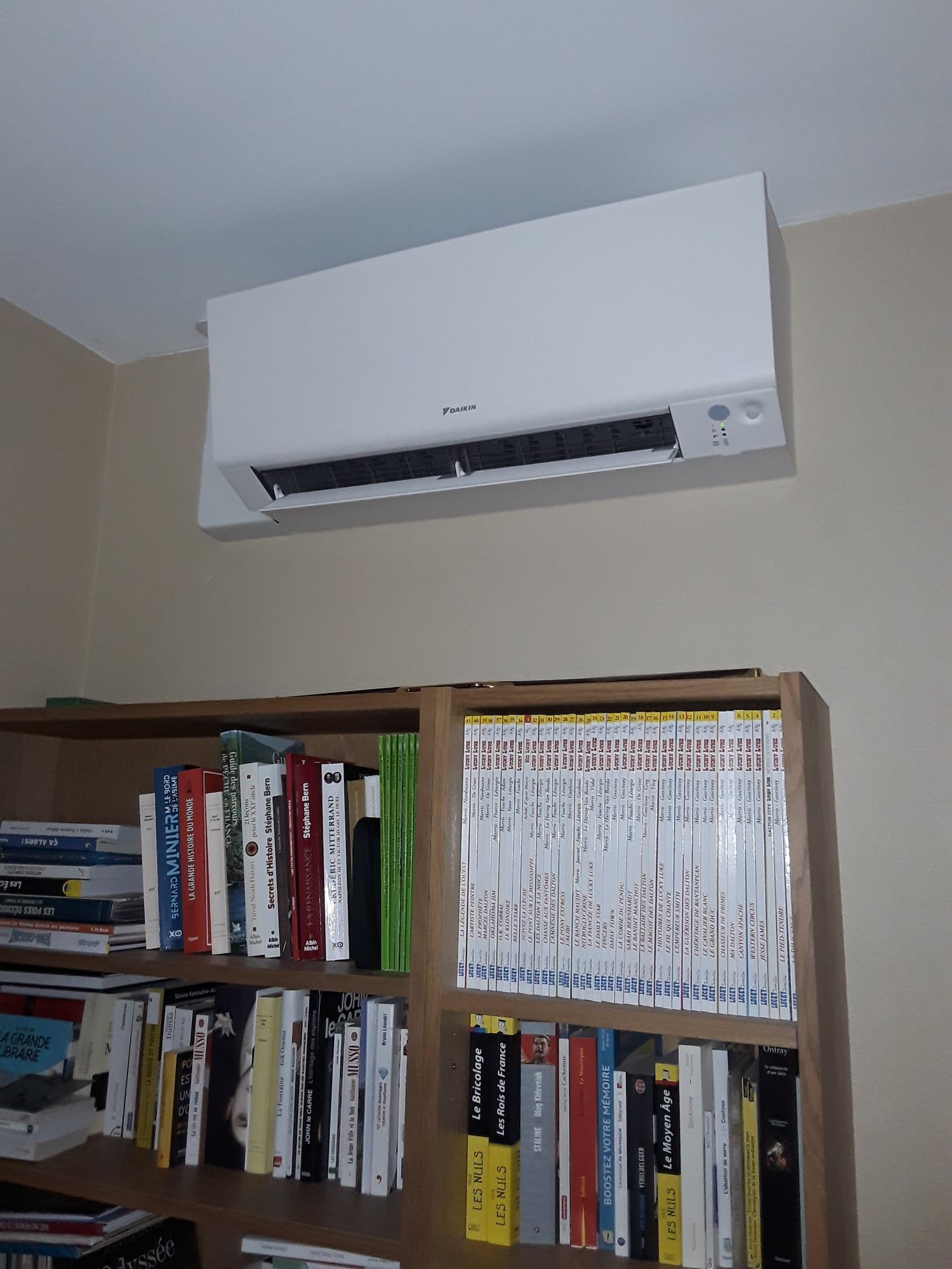 Unité de climatisation fixée au-dessus d'une bibliothèque remplie de livres, contre un mur beige.