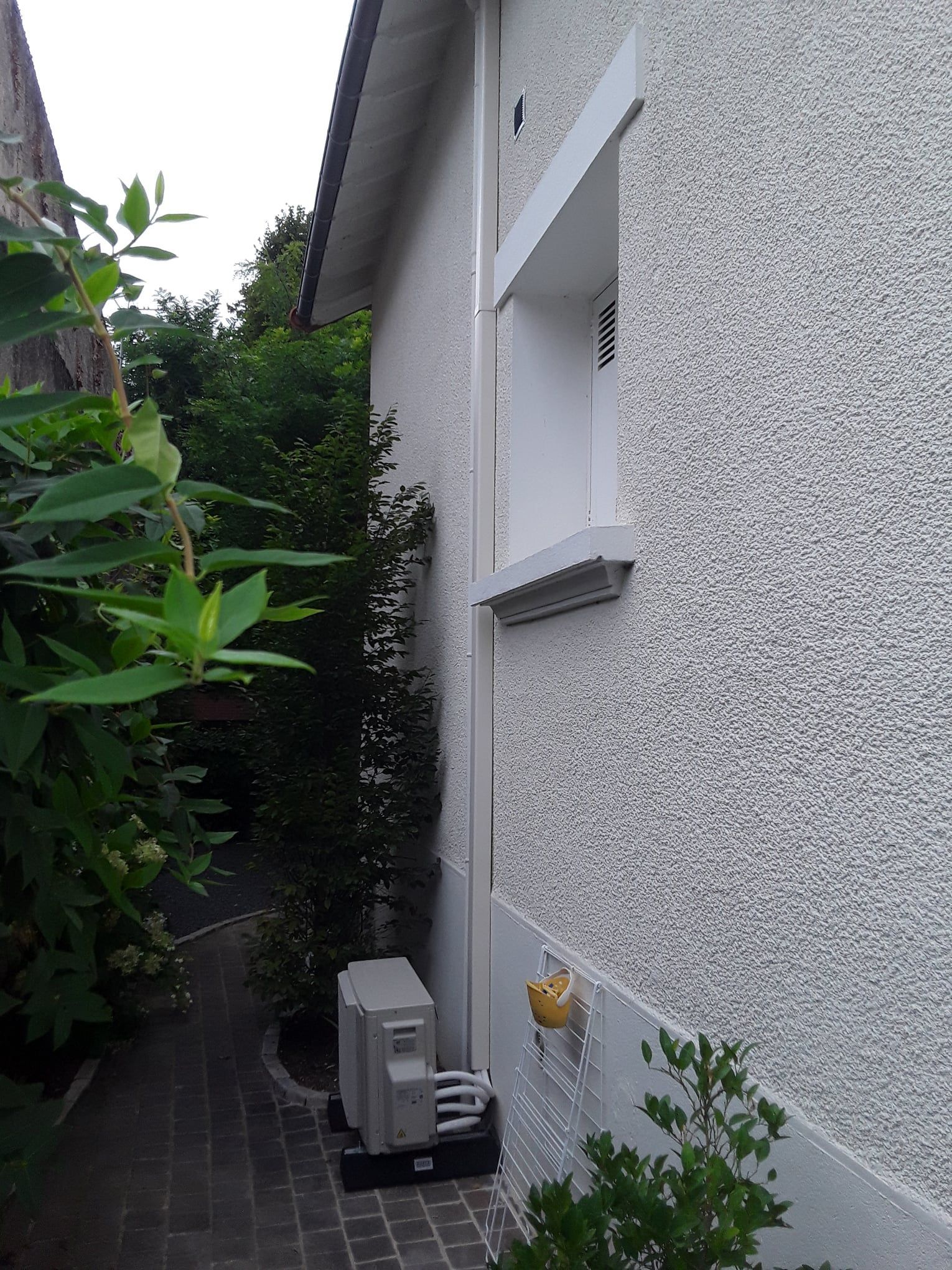 Côté d'un bâtiment blanc avec un enduit texturé, une allée étroite et un climatiseur.