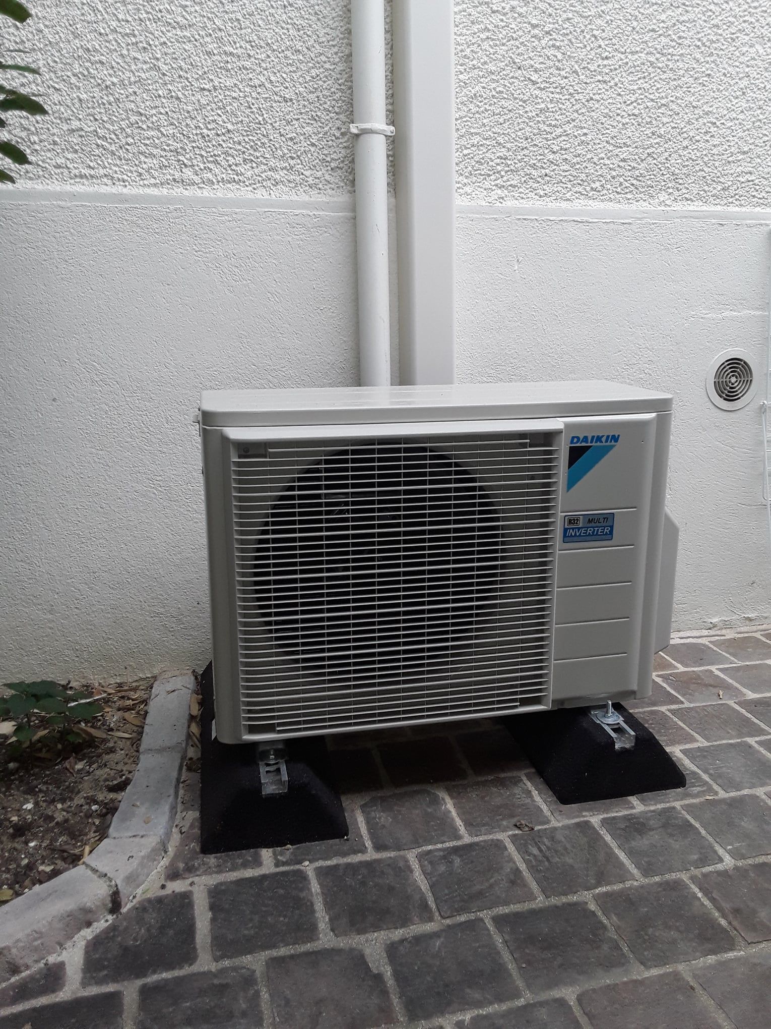 Unité de climatisation extérieure, bleu clair et argent, sur des blocs noirs, contre un mur blanc.