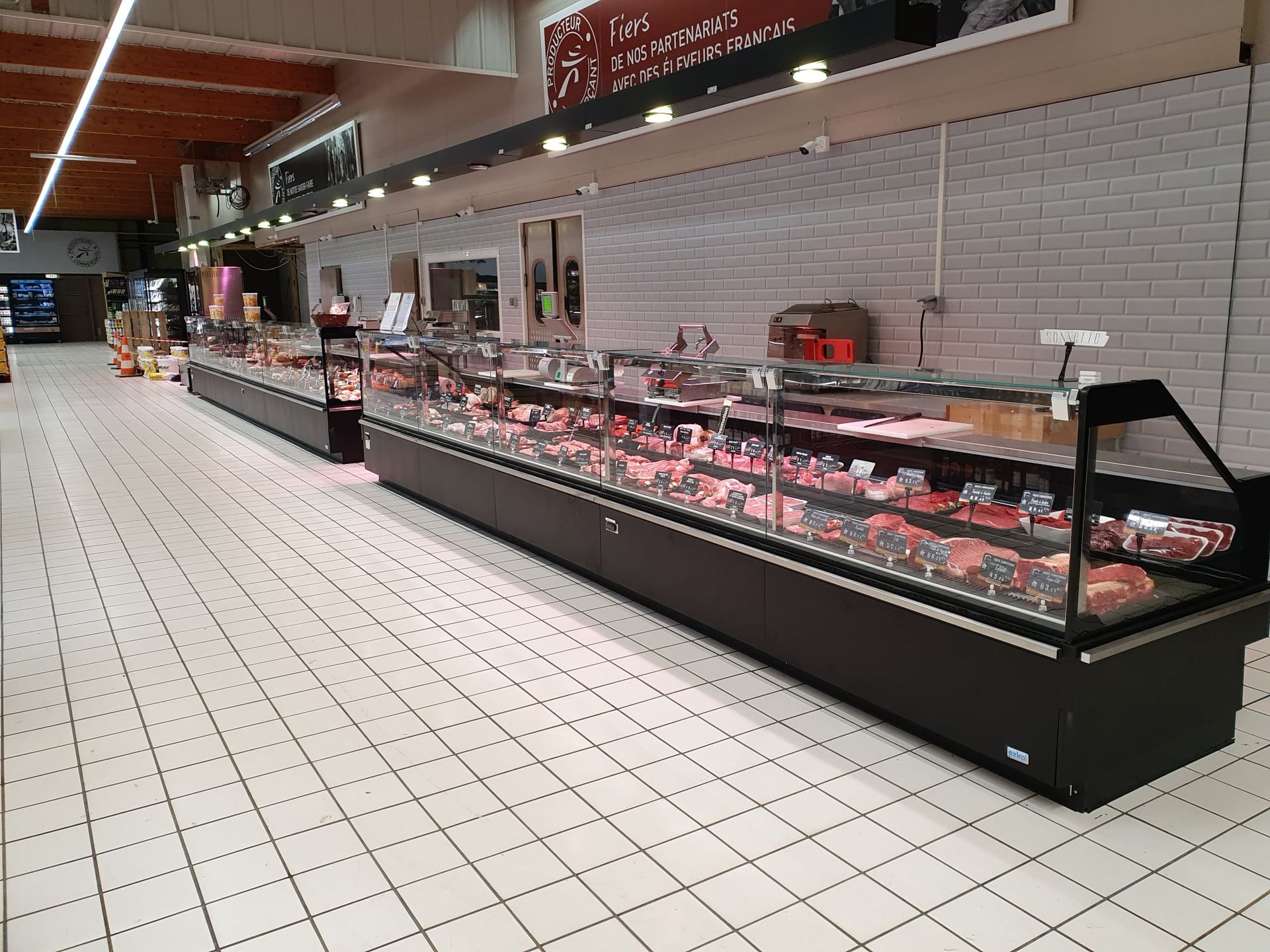 Vitrine à viande dans une épicerie, présentant différentes coupes de viande. Socle noir, sol carrelé blanc.