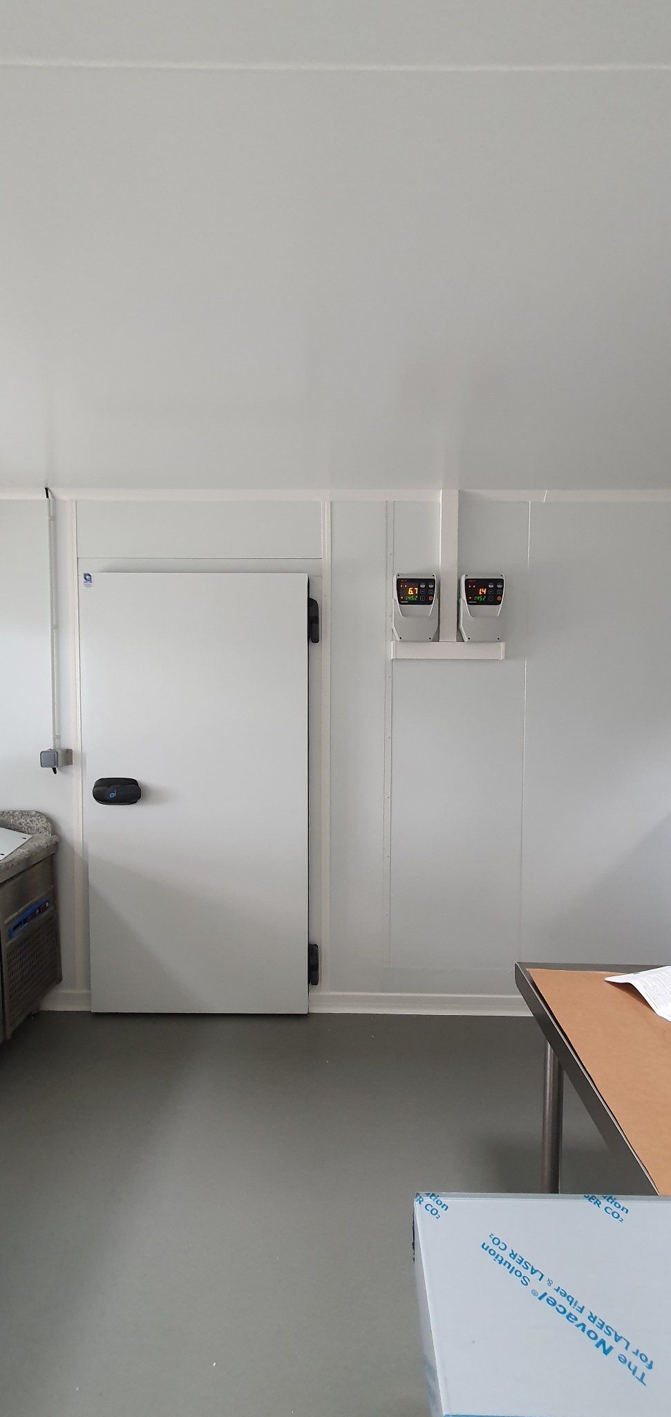 Une pièce blanche avec une porte blanche, un sol gris et deux boîtes noires au mur. Une table et un objet au premier plan.